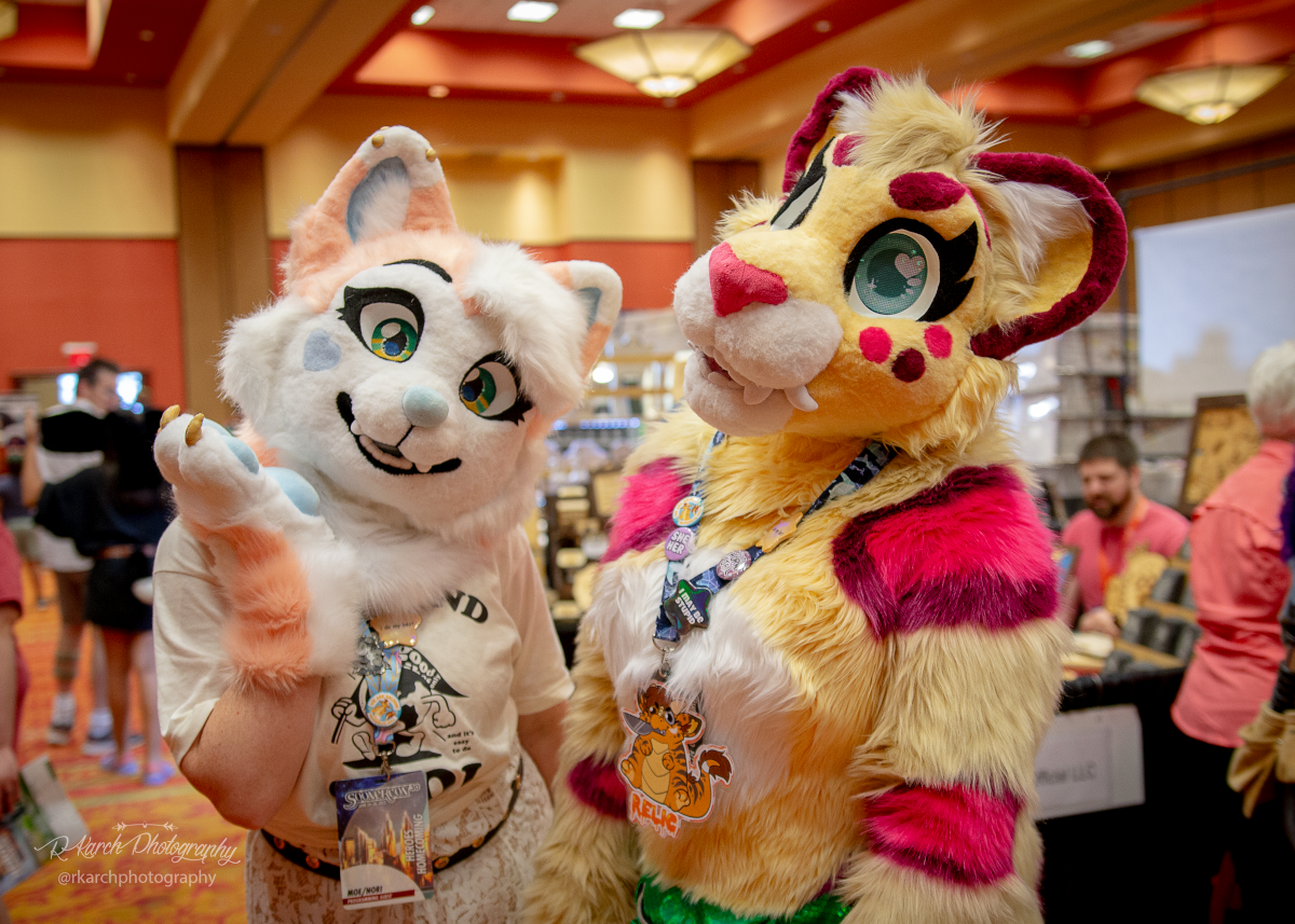 Two costumers in furry costumes pose. One is a white cat and the other is a yellow and pink critter.